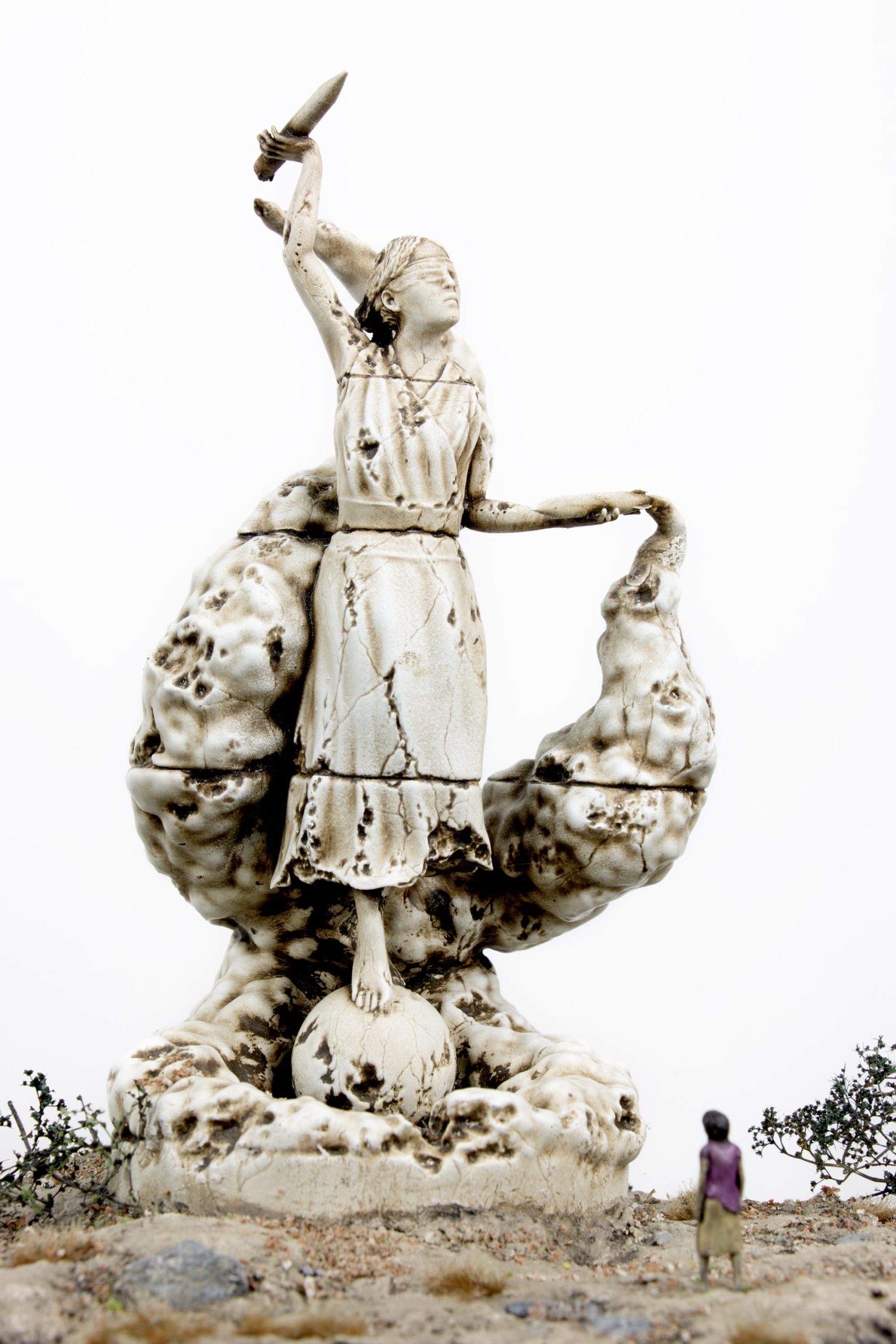 a miniature person sitting at the base of a figurative statue who is blindfolded with a spear in her hand and an explosion coming from her hand