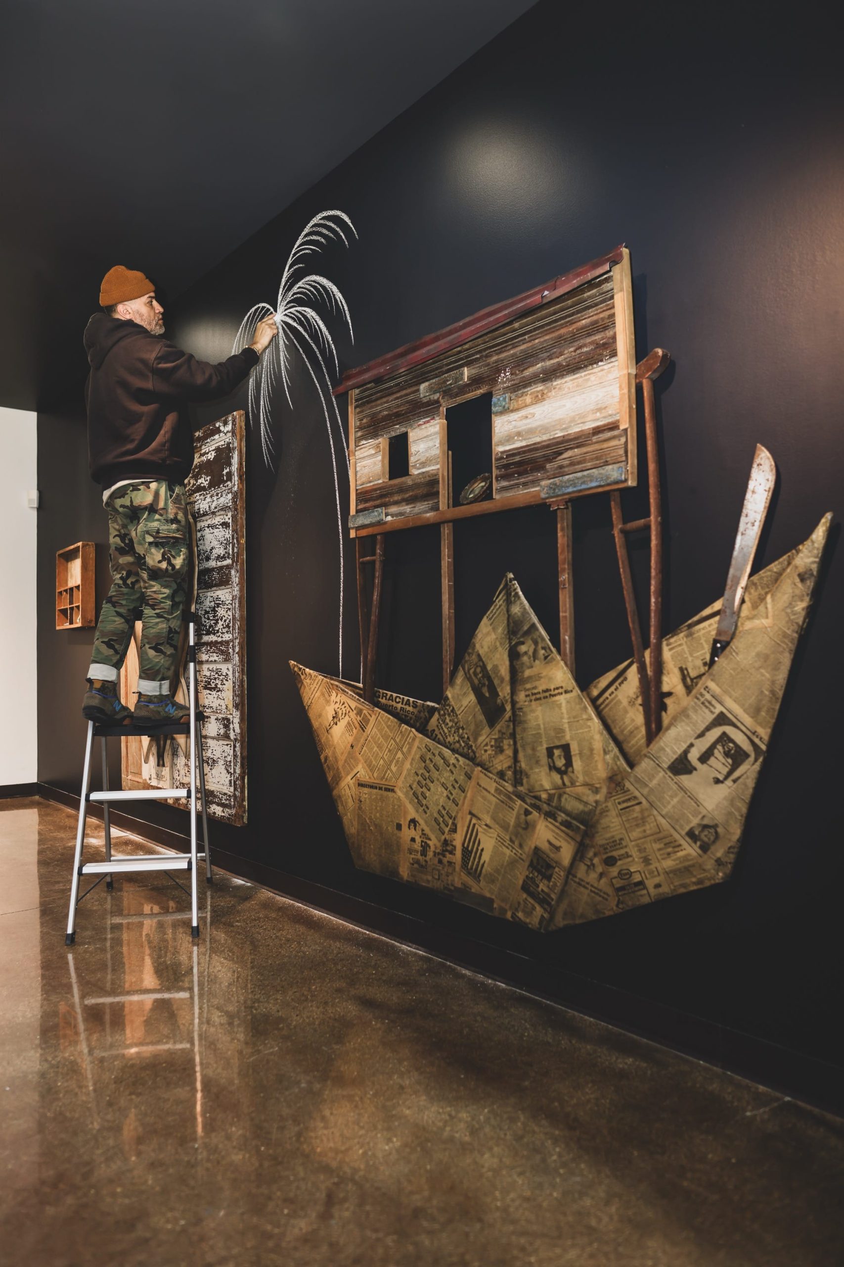 the artist on a ladder installing a feather in a gallery with a large newspaper boat on the wall