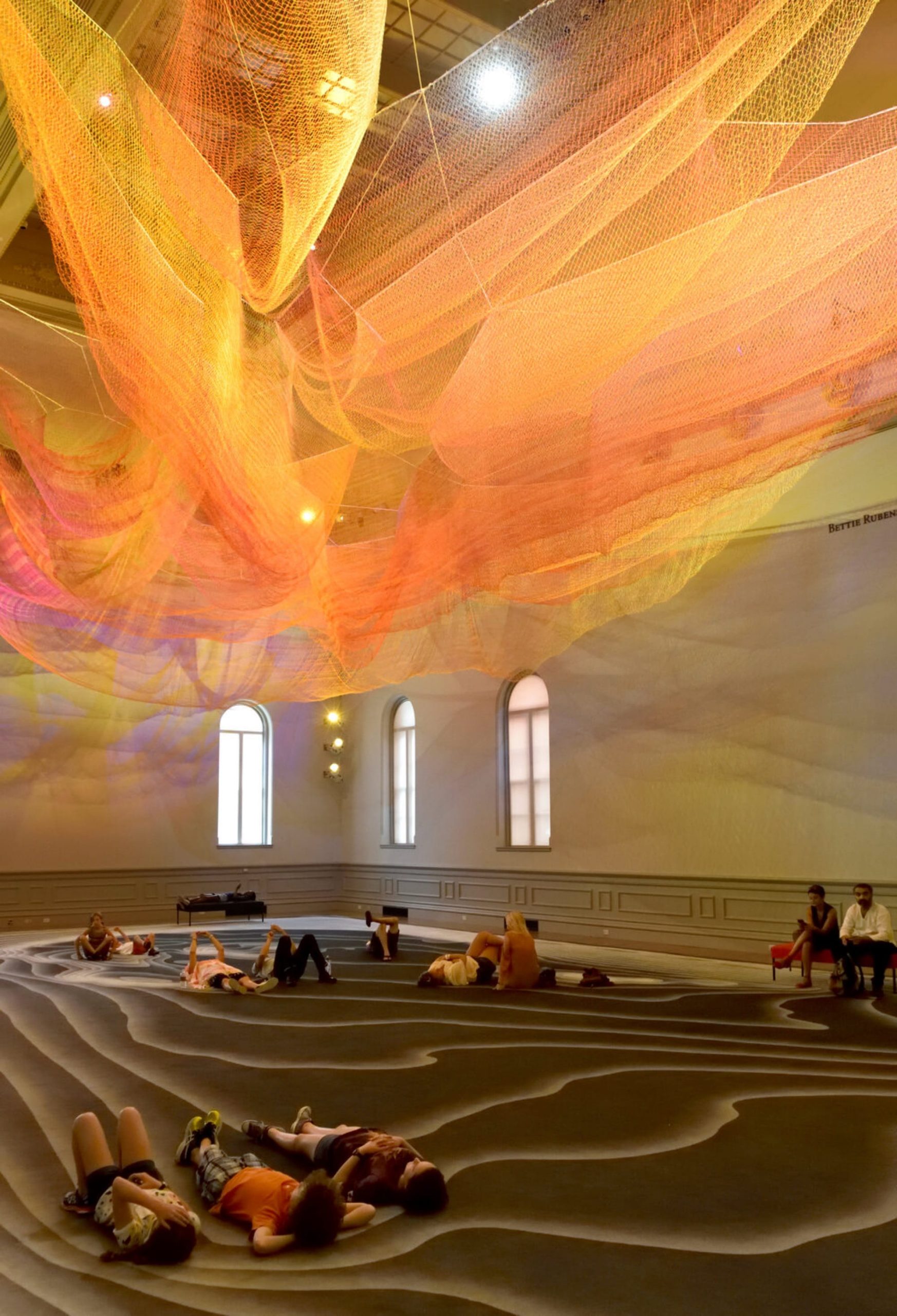 people lay underneath a green and pink Janet Echelman installation suspended from the ceiling