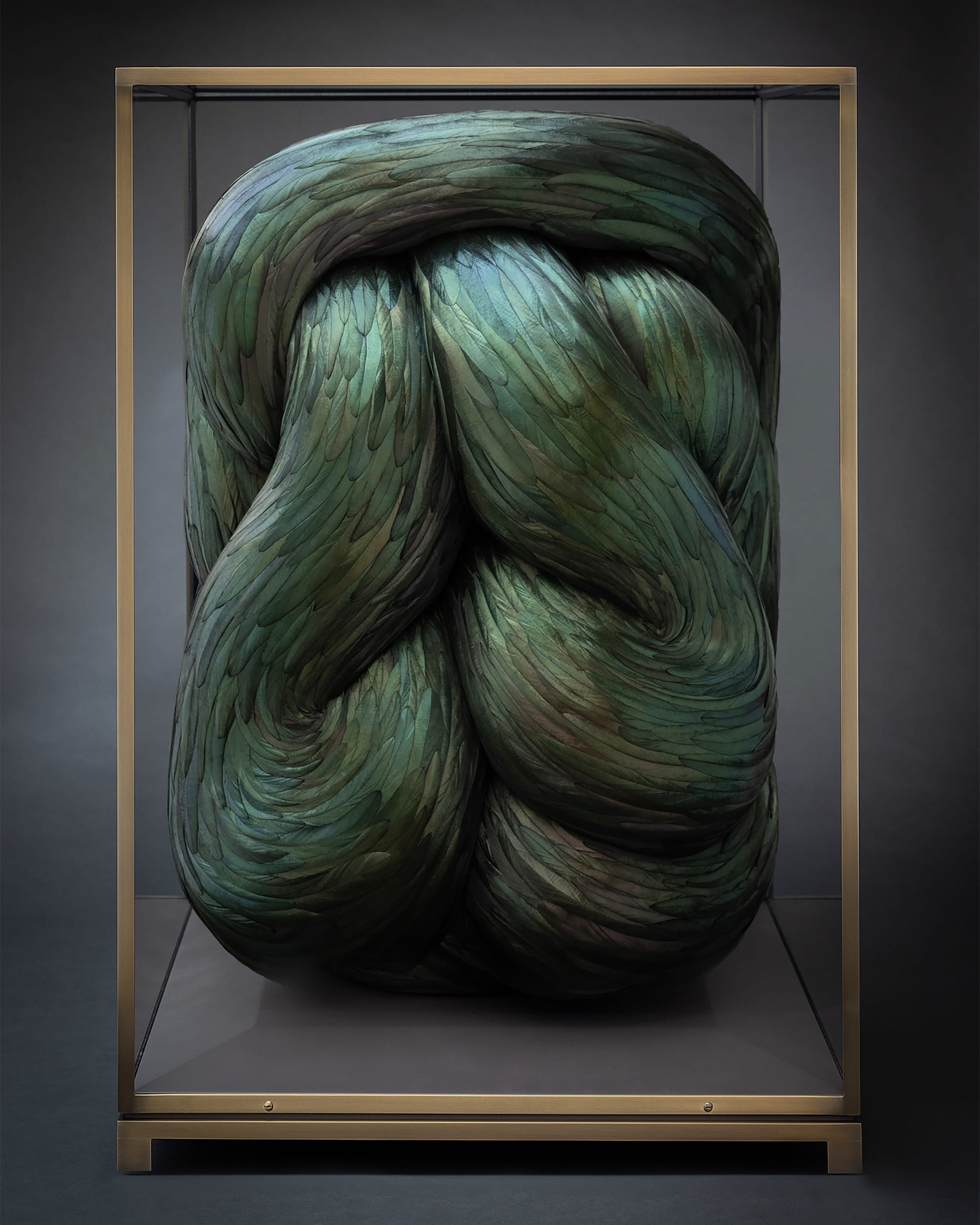a sculpture made of green feathers that appear to be serpentine and knotted inside of a glass vitrine