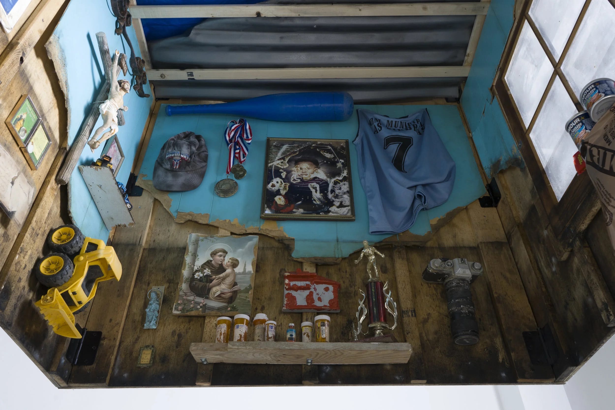 a wooden square structure with found mementos like trophies, jersey, medals, toys, prescription bottles, and religious iconography