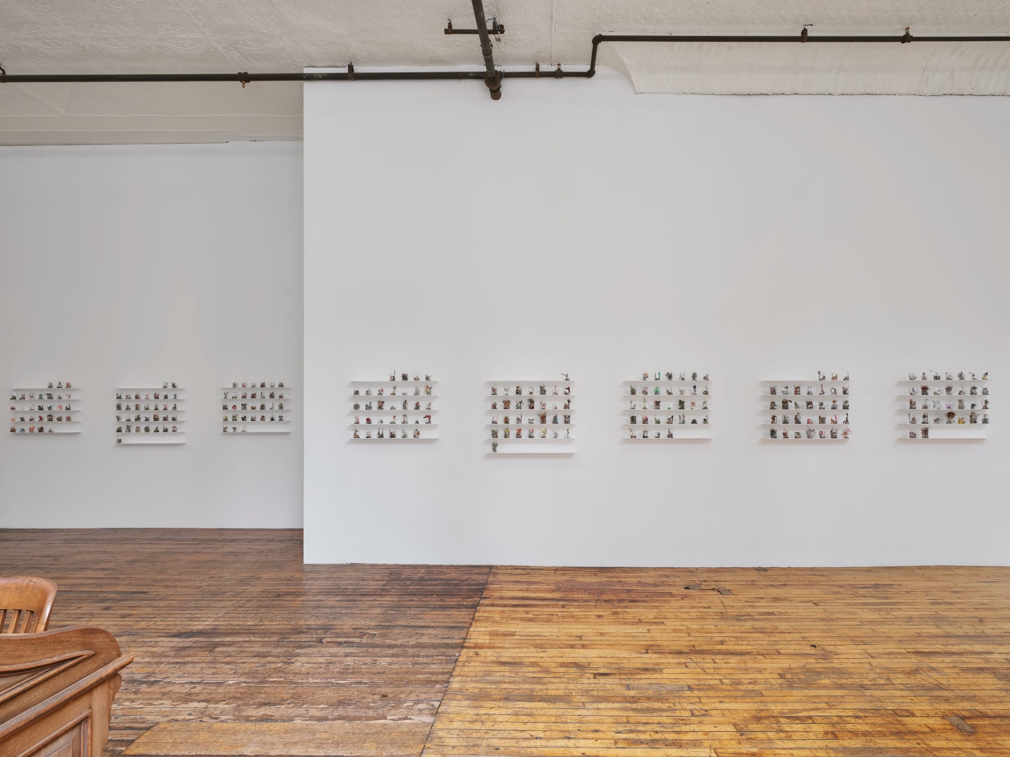 a collection of small ikebana-like sculptures in cellophane arranged like a month calendar on white shelves. several of these collections line a gallery wall with wood floor