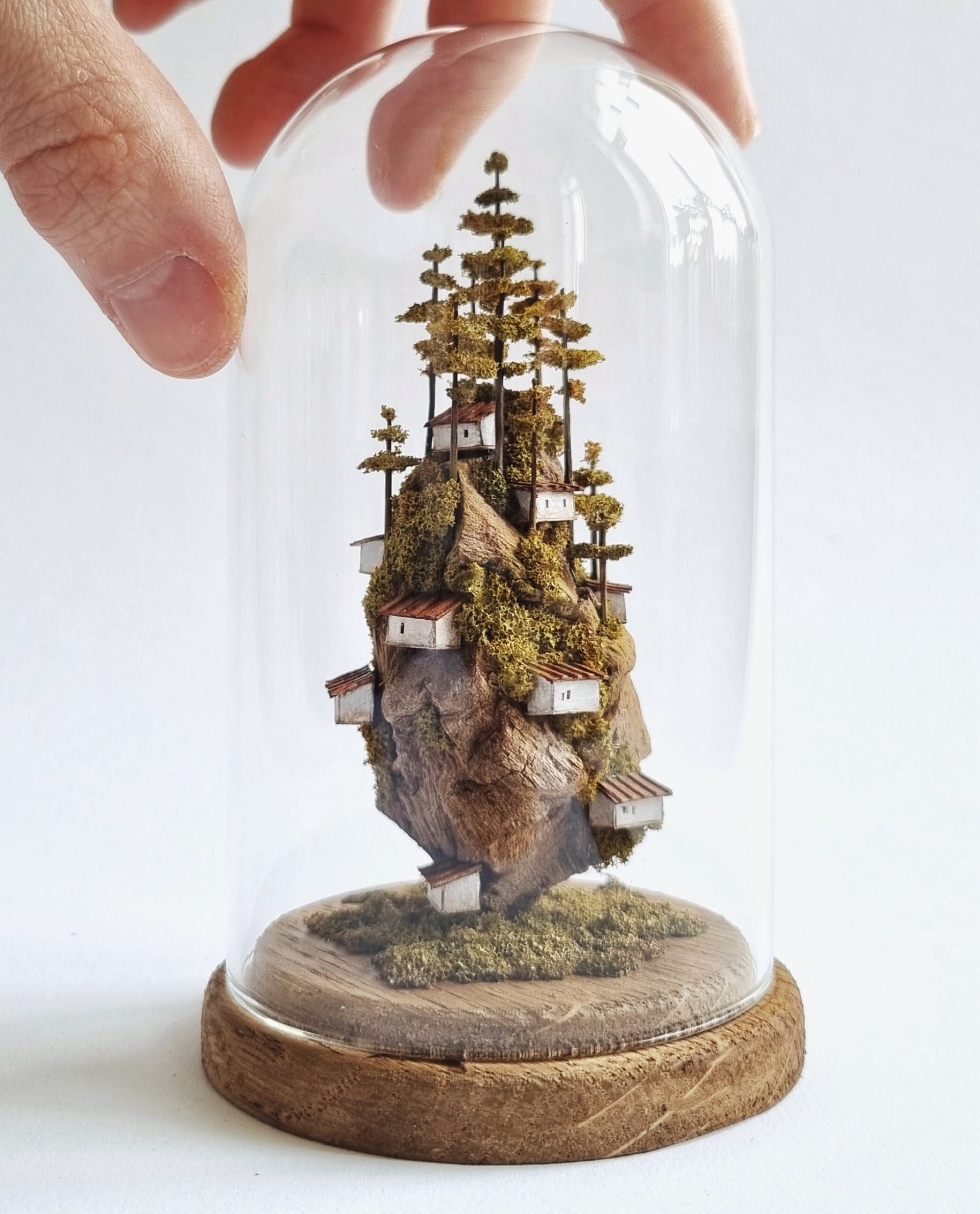 a hand touches a glass dome around a miniature sculpture of tiny, stacked houses and trees by Michael Davydov