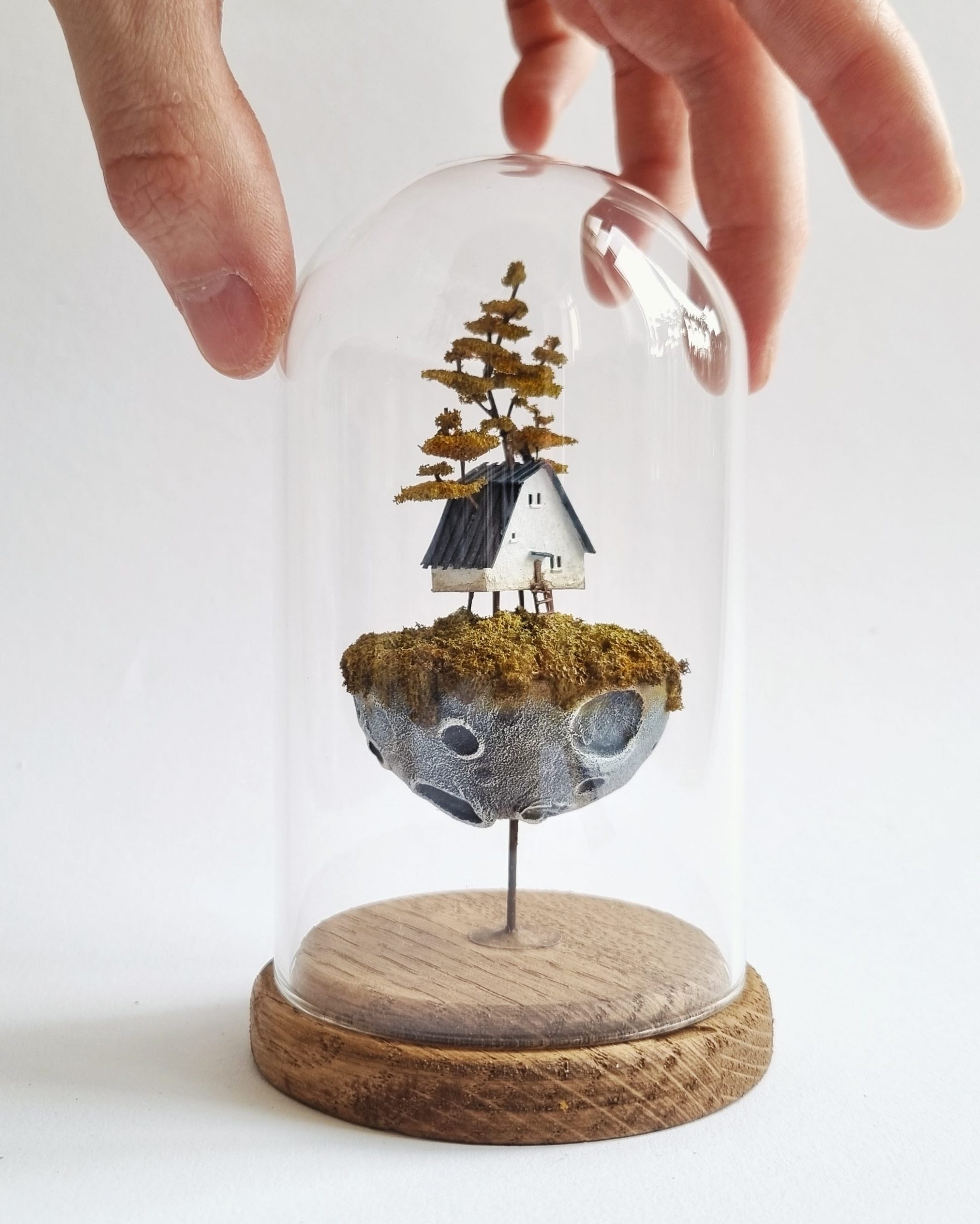 a hand touches a glass dome around a miniature sculpture of tiny, stacked houses by Michael Davydov