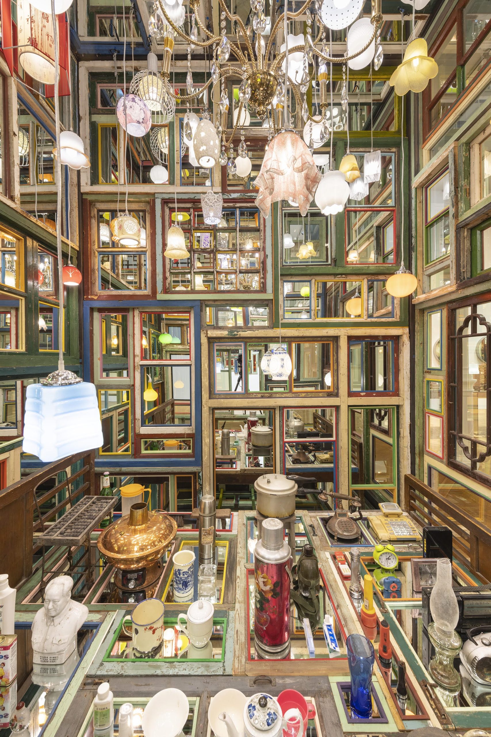 an interior detail of an installation by Song Dong using found wooden window frames, color-coated glass, lamps, and other knick-knacks