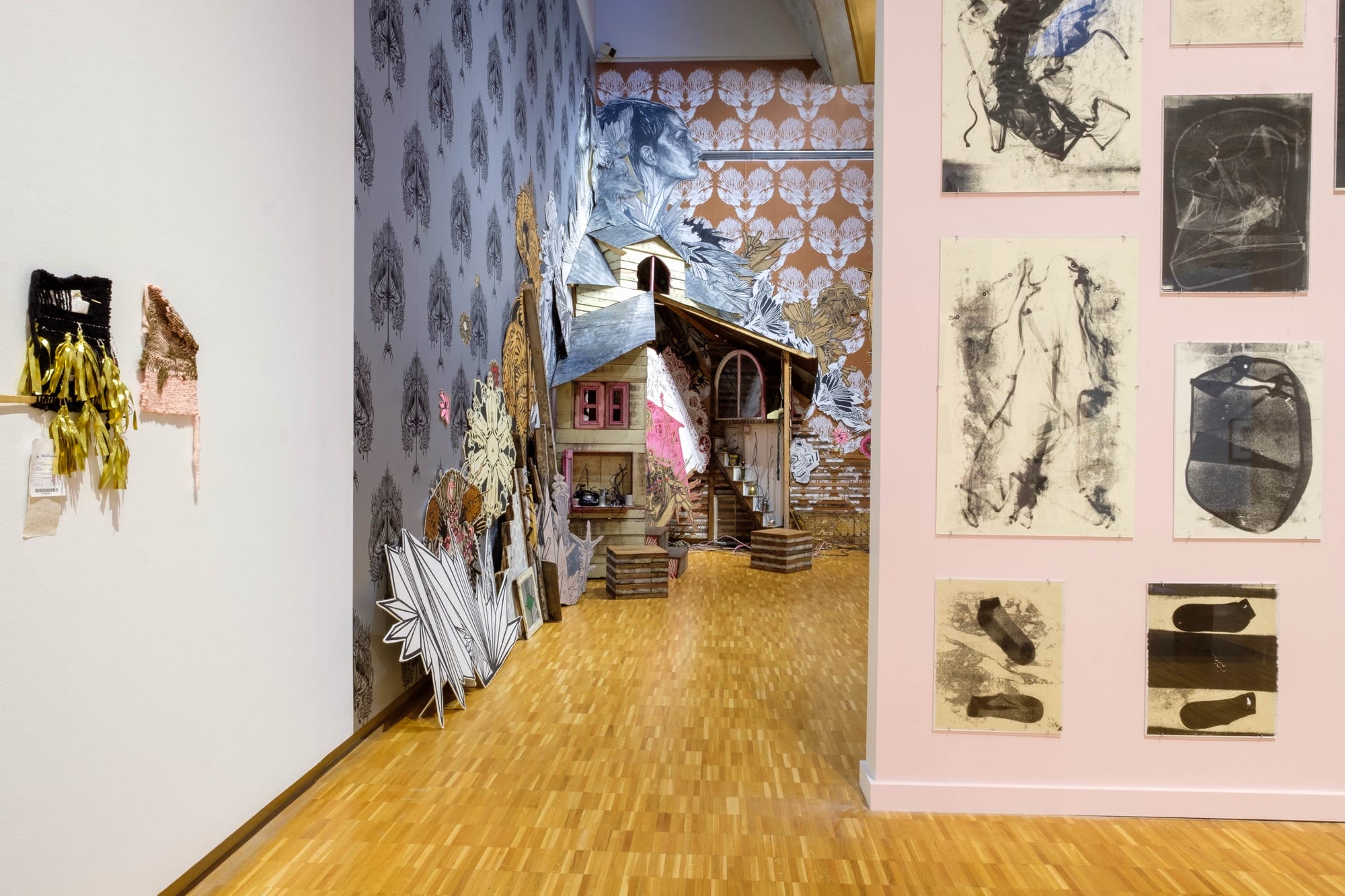 in the background is an an installation by Swoon with patterned wallpaper, figures, vignettes, and a tarantula woman at the center. a pink wall with monoprints and small weavings by Bryana Bibbsare in the foreground. the pieces are in a gallery at the Haggerty Museum of Art.