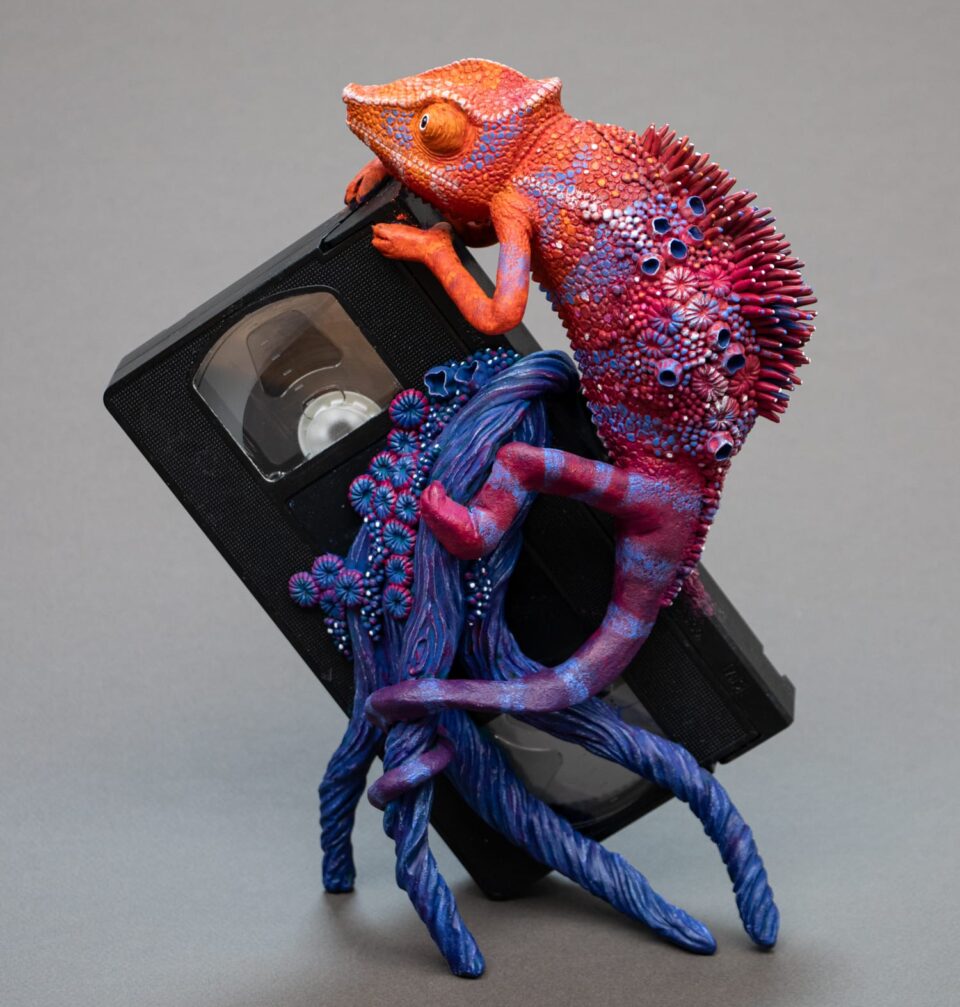 a sculpture by Stéphanie Kilgast of a colorful chameleon climbing on a VHS tape